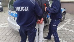 Policjanci umieszczają zatrzymanego w radiowozie oznakowanym. Policjanci umundurowani prowadzą zatrzymanego, który ma założone kajdanki na ręce trzymane z tyłu.