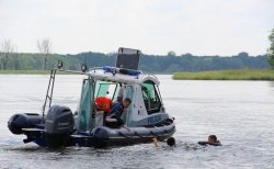 Zdjęcie przedstawia łódź motorową policji znajdująca się na terenie zbiornika wodnego i policjanta udzielającego pomocy dwóm osobom znajdującym się w wodzie