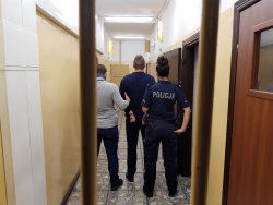 Zatrzymany sprawca pobicia mieszkańców Krajenki stoi tyłem trzymany przez policjantów w pomieszczeniu dla osób zatrzymanych