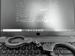Komputer przenośny na jego klawiaturze leżą policyjne kajdanki, na ekranie zaś widać ciąg różnych znaków i napis hacking