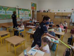 Dwie umundurowane policjantki wręczają uczniom klas pierwszych elementy odblaskowe. Dzieci siedzą w ławkach szkolnych