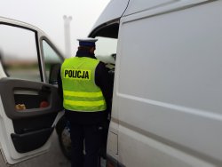 Policjant prowadzi kontrolę uczestnika ruchu drogowego. Widoczne otwarte drzwi pojazdu od strony kierowcy i policjanta odwróconego tyłem
