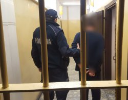 Zdjęcie przedstawia policjanta w pomieszczeniu dla osób zatrzymanych, który prowadzi zatrzymanego mężczyznę do pokoju dla osób zatrzymanych mężczyzna ma założone kajdanki na ręce trzymane z tyłu